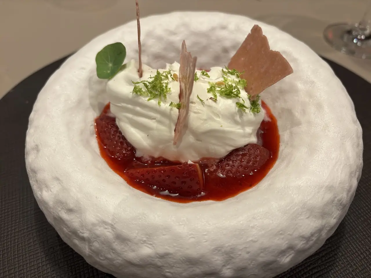 A white cream dessert topped with herbs and a thin wafer, set in a pool of red sauce in a textured white bowl