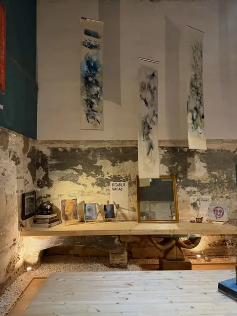 Rustic cafe interior with distressed plaster-and-stone walls, hanging abstract watercolour scrolls and a wooden shelf displaying framed artwork and a small vitrine