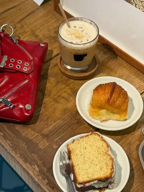 An iced coffee, a croissant on a small plate and a slice of glazed lemon cake on a wooden table, next to a red leather bag