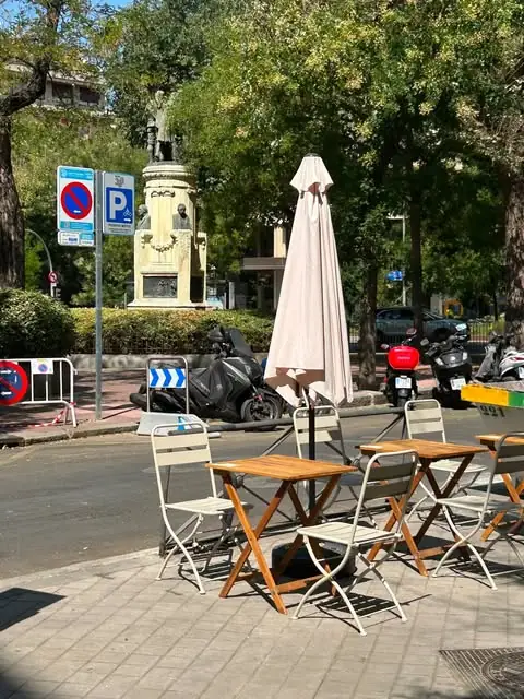 Sidewalk terrace with wooden folding tables and white metal chairs under a closed parasol, a stone monument, trees and parked scooters across the street