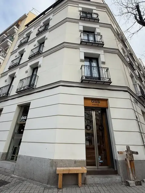 Exterior corner storefront of OSS KAFEE in a cream-coloured building with wrought-iron balconies, a glass door and a small wooden bench outside