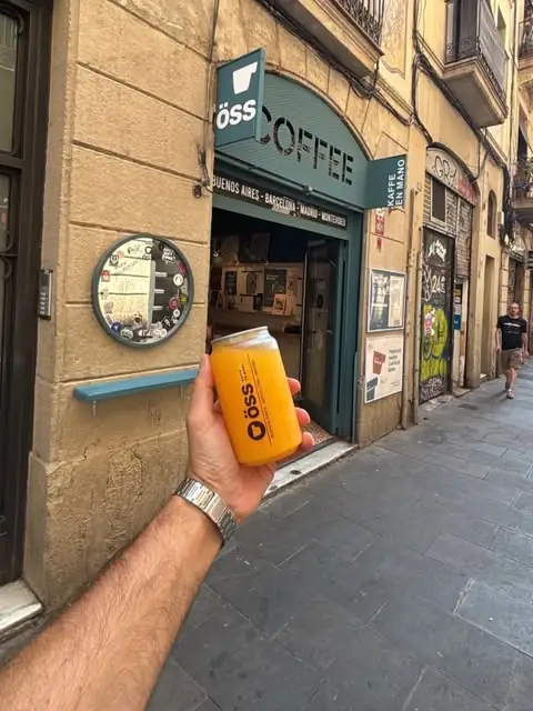 Hand holding an OSS-branded orange drink can in front of the OSS COFFEE storefront, whose green sign lists Buenos Aires, Barcelona, Madrid, Montevideo
