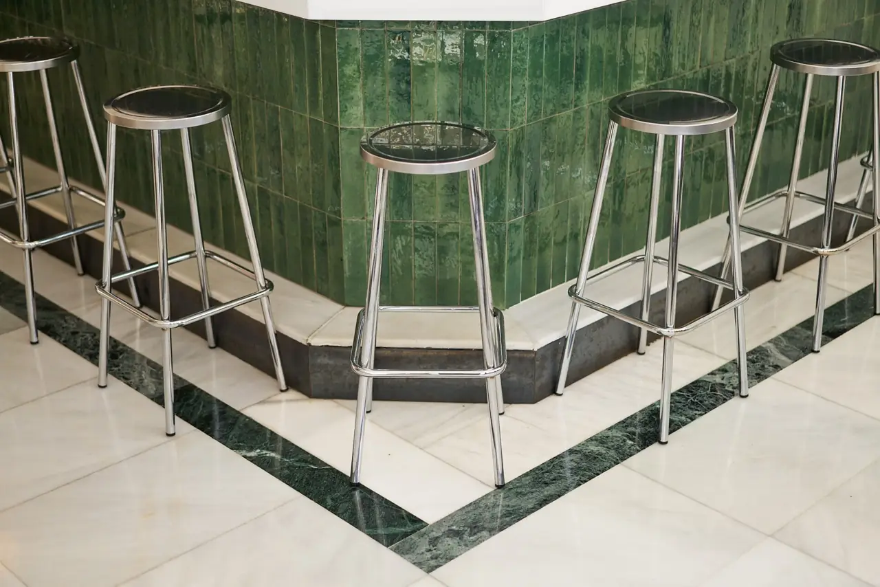 Interior detail of chrome bar stools around a green-tiled bar base with white and green marble flooring