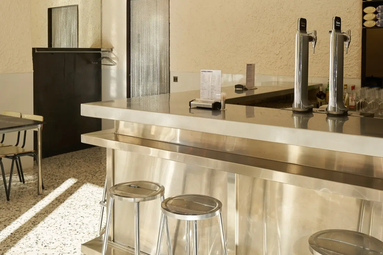 Stainless-steel bar with two beer taps, bottles and a menu card reading OSTRAS PEDRIN, with metal stools and a dining table alongside