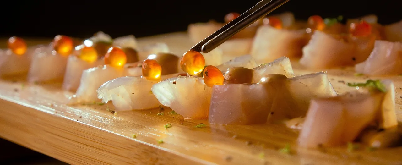 Slices of raw white fish topped with orange roe on a wooden board with tweezers
