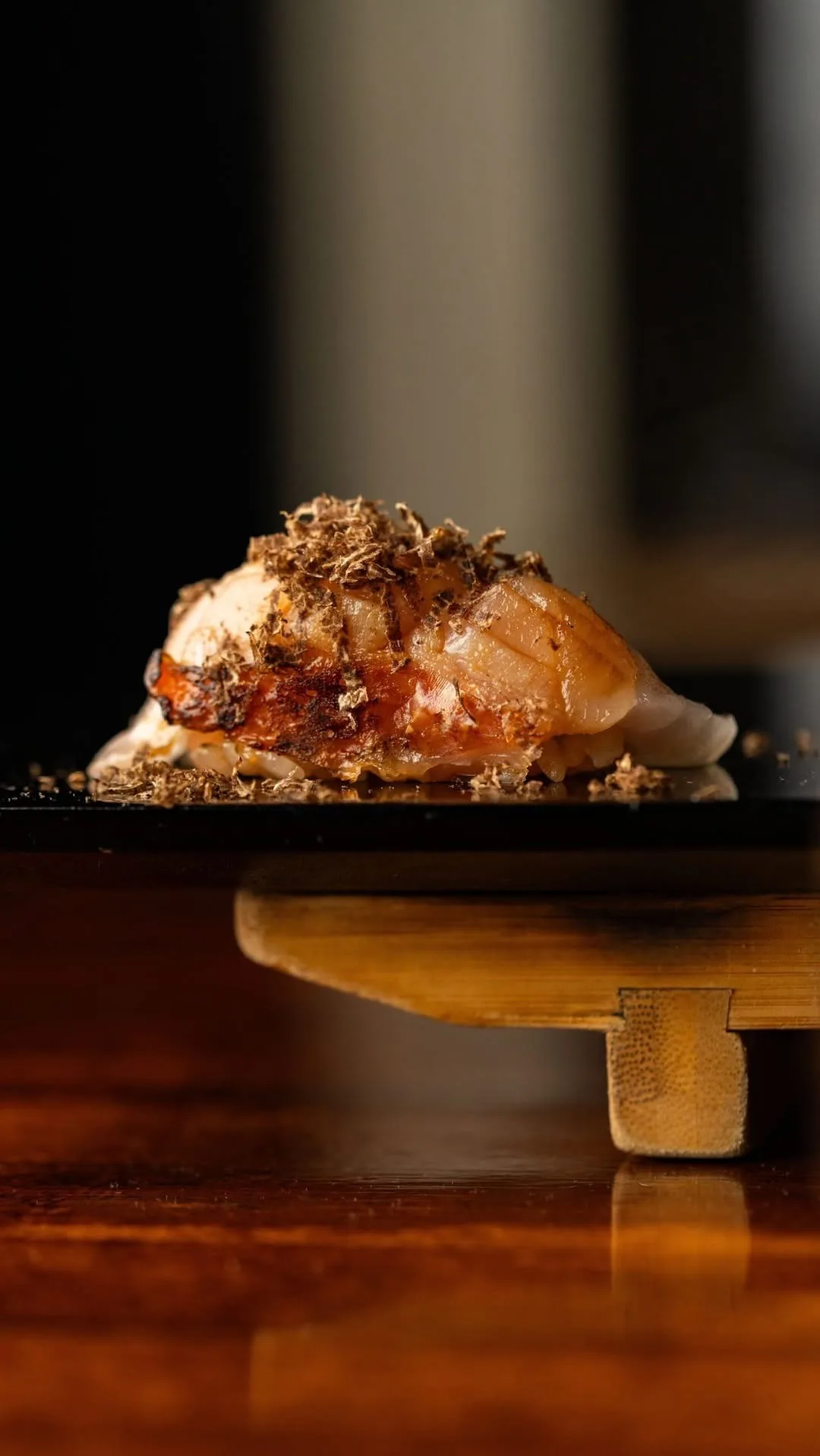 A nigiri topped with browned fish and shaved truffle on a black tray