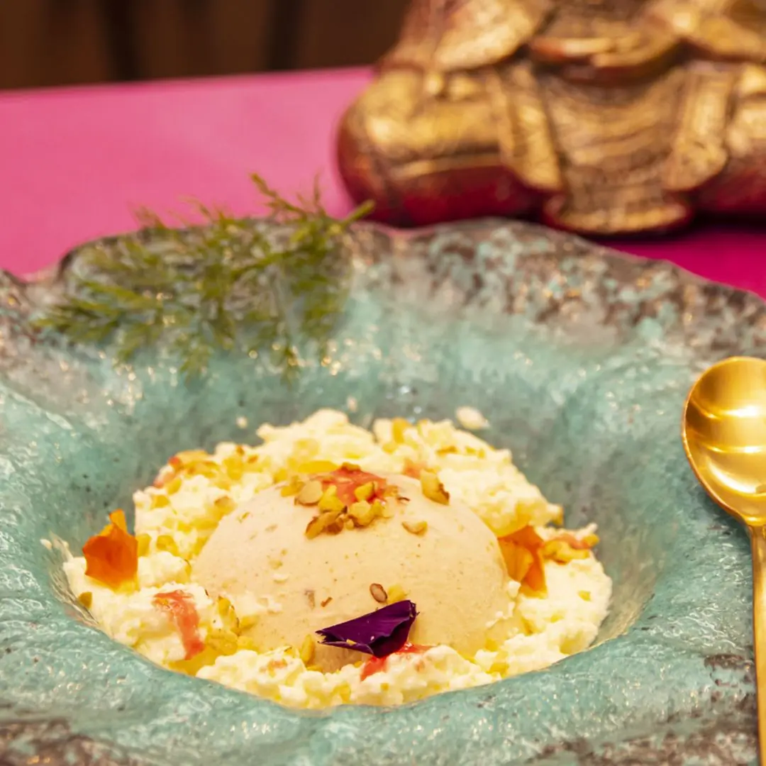 A dome-shaped ice cream dessert on a bed of shredded coconut, topped with crushed pistachios and a rose petal, in a textured ceramic bowl, at Out of India