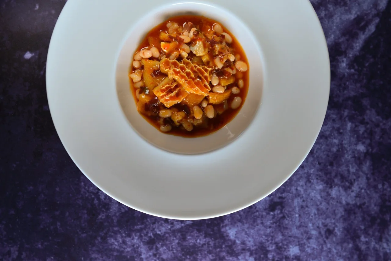 A top-down view of a white bowl of tripe and white beans in a red sauce on a purple background