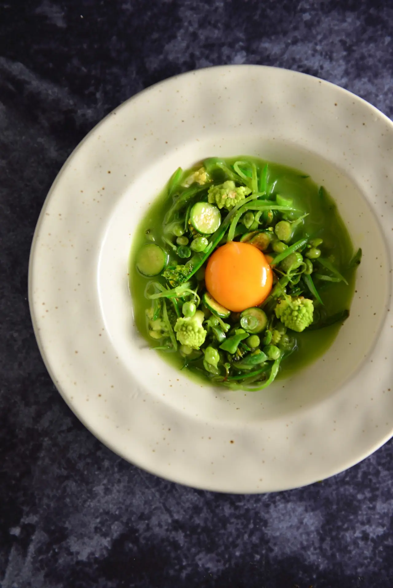 Overhead view of a speckled cream bowl holding green vegetables - romanesco florets, peas, broad beans, courgette slices and shredded greens - in a bright green broth around a raw orange egg yolk