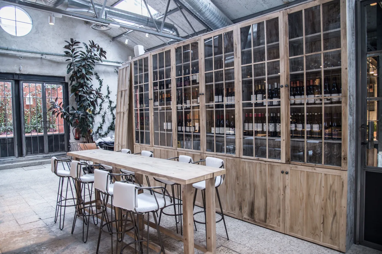 A long wooden table with high stools in front of a large glass-fronted wine cabinet and a leafy plant