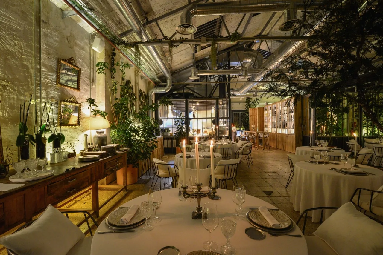A candlelit dining room with round white-clothed tables, plants, a high glass-paned wall and a wooden wine cabinet