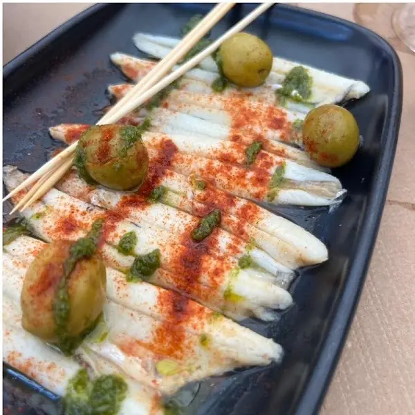 Grilled small fish fillets drizzled with green herb sauce and paprika, garnished with whole olives on a black plate, at Oviso.