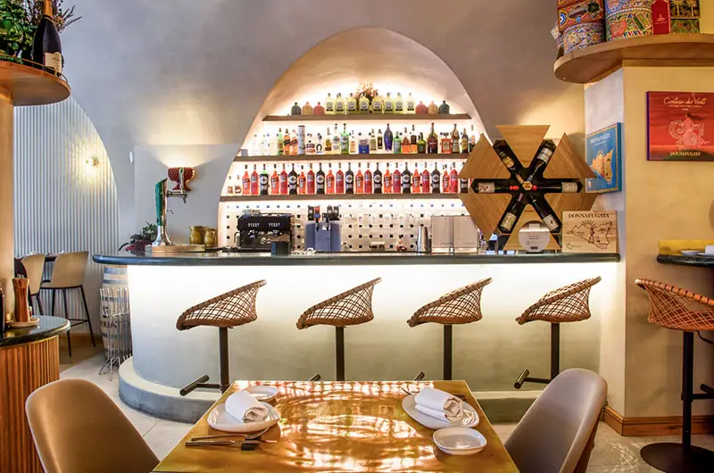 Restaurant bar with an arched niche of backlit liquor bottles, rattan bar stools and a set wooden table in the foreground