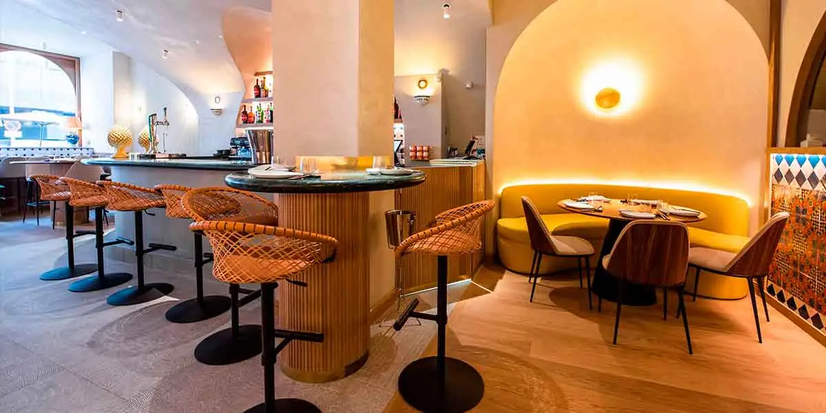 Bright dining room with a curved bar, rattan bar stools, a yellow curved banquette and a set table under a wall light