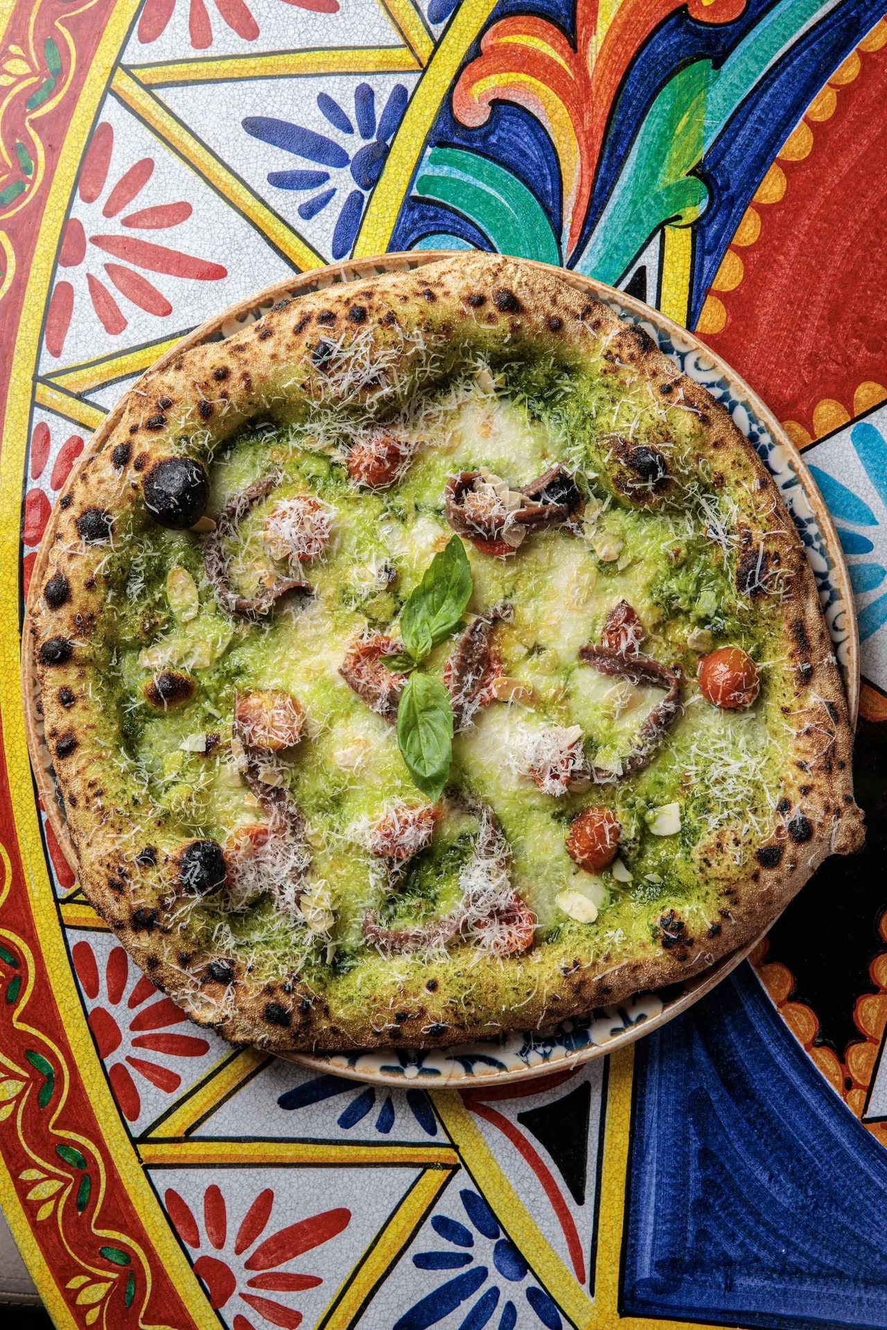 A pizza with green pesto, red tomato and basil on a colourful patterned tray