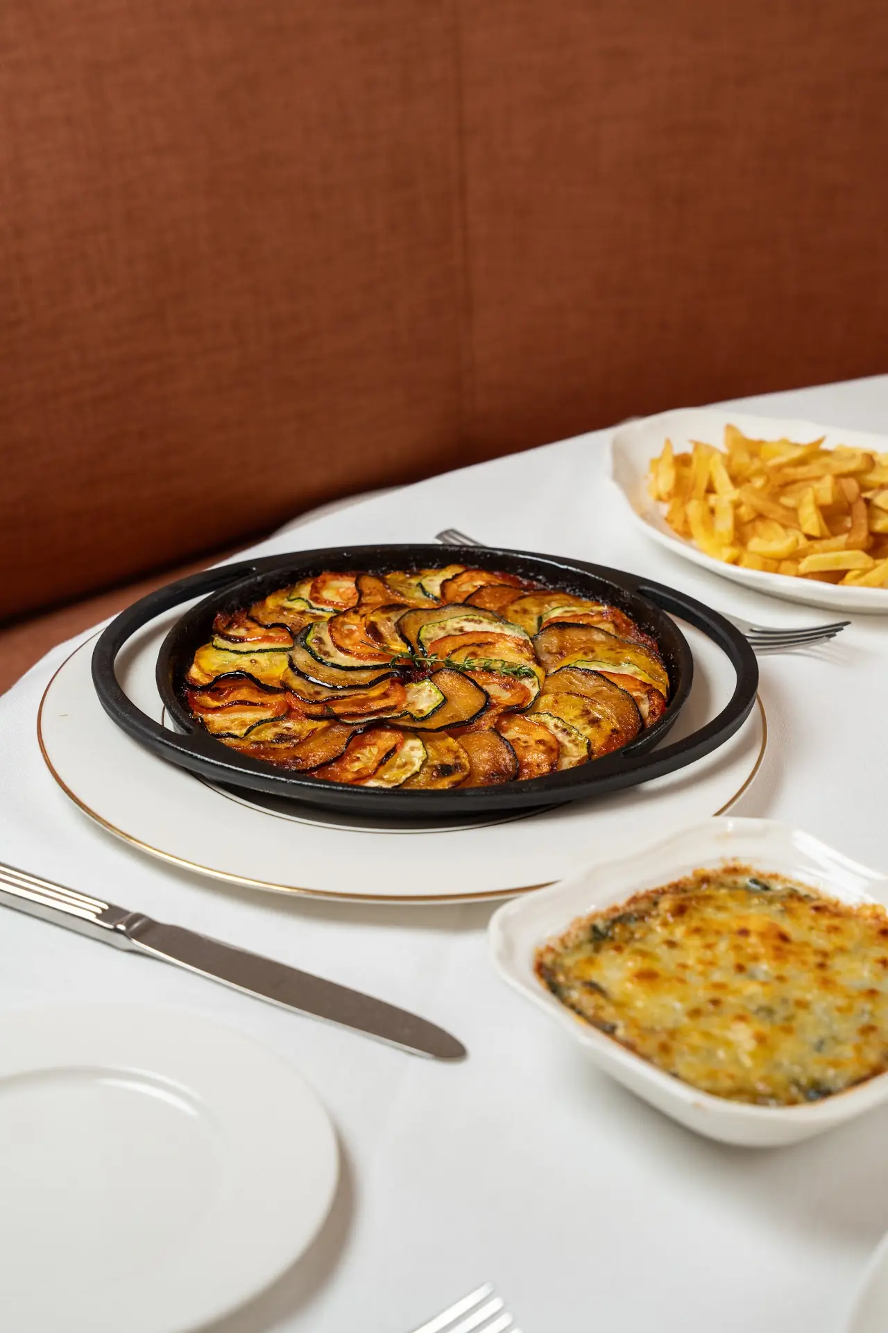 Sliced vegetable gratin in a black dish alongside fries and a cheese gratin