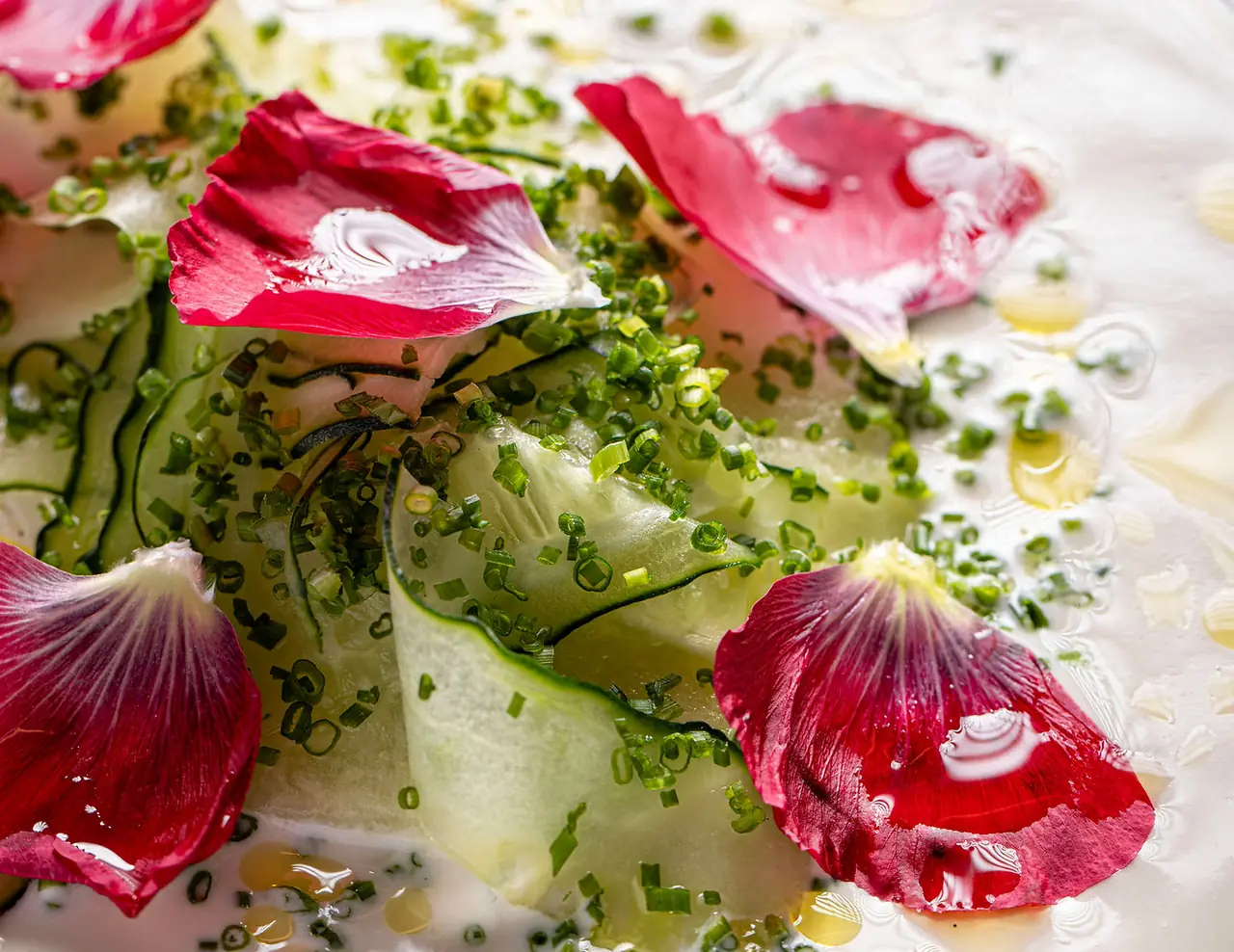 Close-up of folded cucumber ribbons scattered with finely chopped chives and topped with deep pink flower petals, sitting in a pale broth beaded with oil