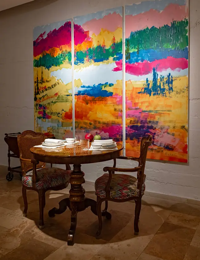 Small round antique table laid with white plates, a pomegranate and a green apple, flanked by two cane-backed armchairs in patterned upholstery, beneath a large three-panel abstract painting in pink, yellow and blue