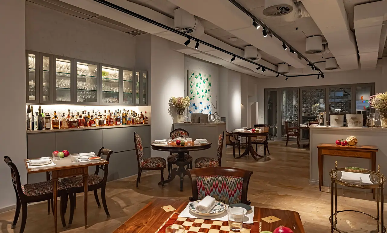 Wide view of the dining room: antique wooden tables laid on a travertine floor with patterned upholstered chairs, a lit back bar of spirits under glass-fronted cabinets of stemware, track lighting on a concrete ceiling