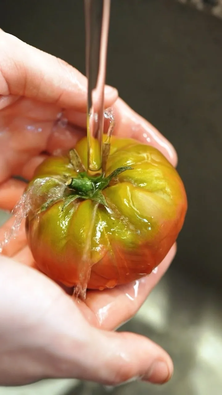A ripe tomato being rinsed under running water in a hand