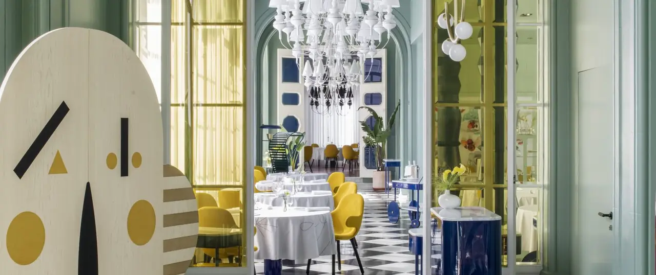 Dining room in mint-green panelling with round tables in white linen, mustard-yellow armchairs, a black-and-white checkerboard floor and a large white ceramic chandelier