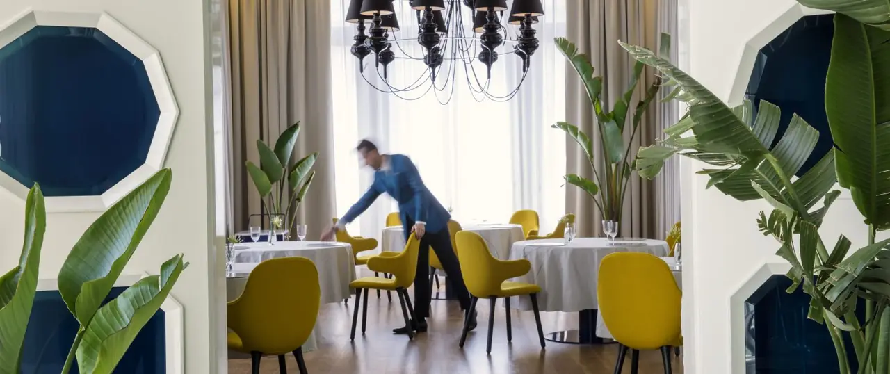 Waiter in a blue jacket, blurred mid-motion, laying a round white-clothed table between mustard-yellow chairs, banana plants and a black chandelier against a bright curtained window