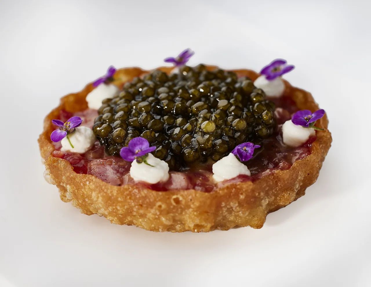 A round crisp tart topped with a mound of dark caviar, small white dollops and purple flowers
