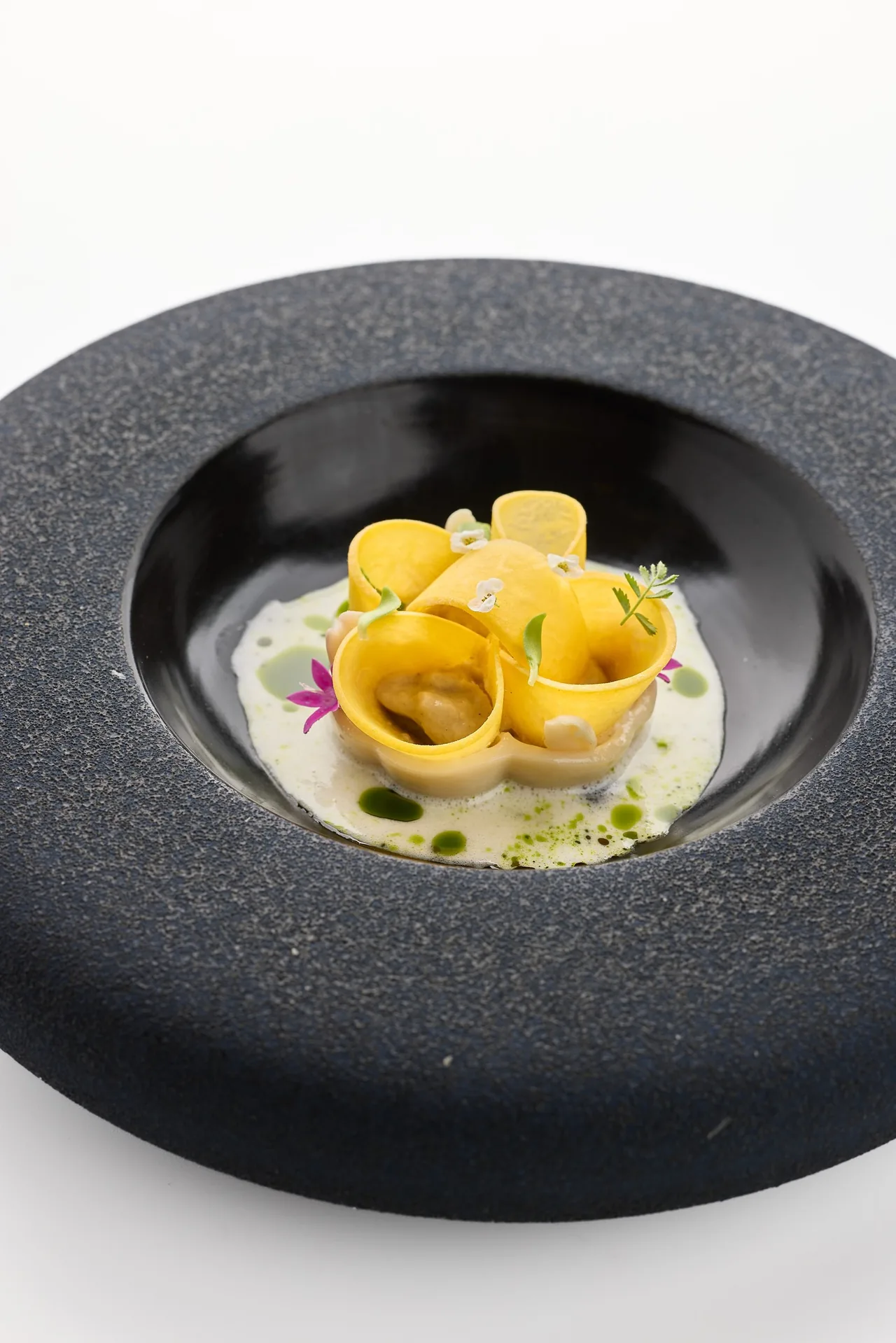 A dark textured bowl holding rolled yellow pasta in a pale sauce with small flowers