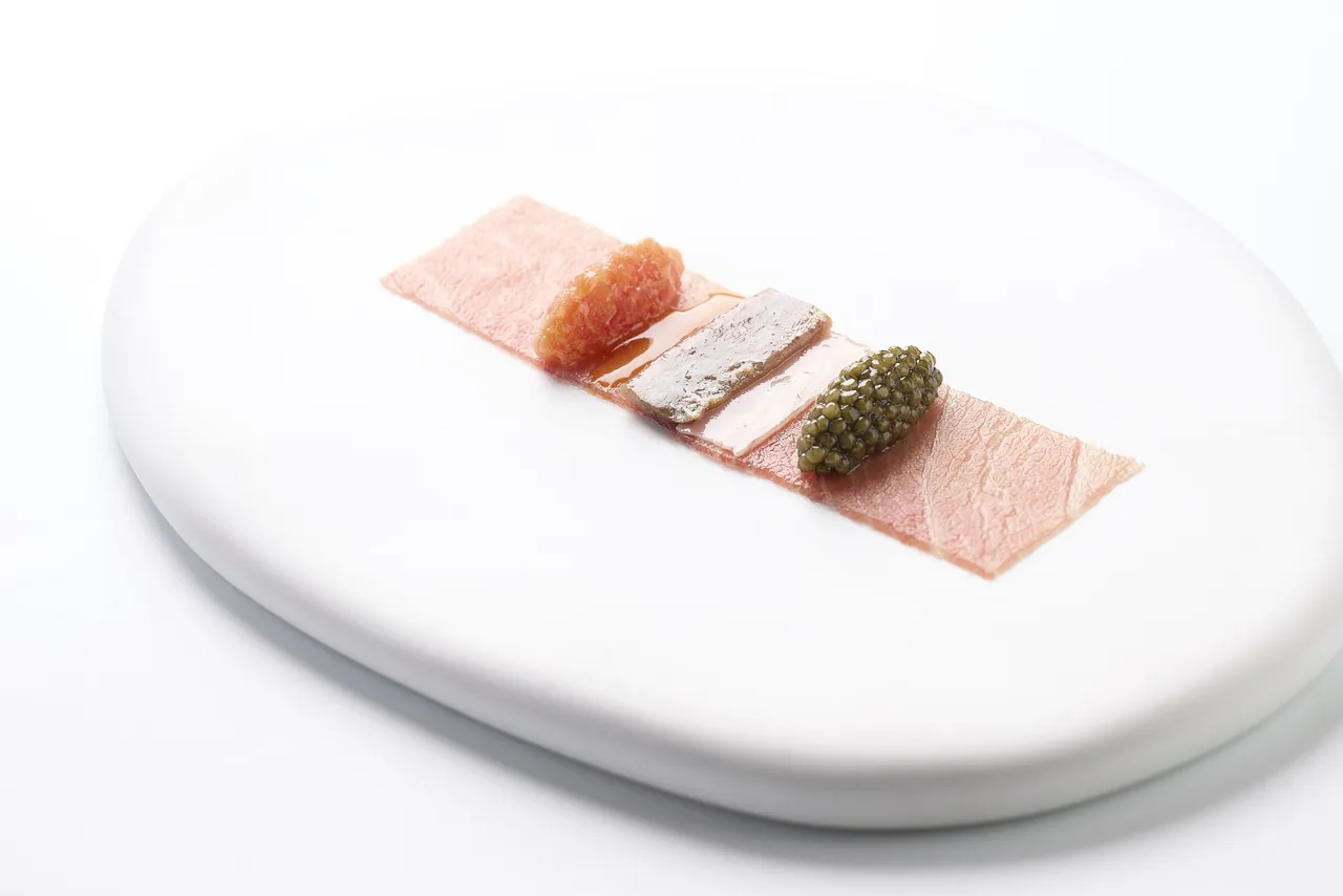 A long white plate with thin slices of pink fish and a small dark garnish