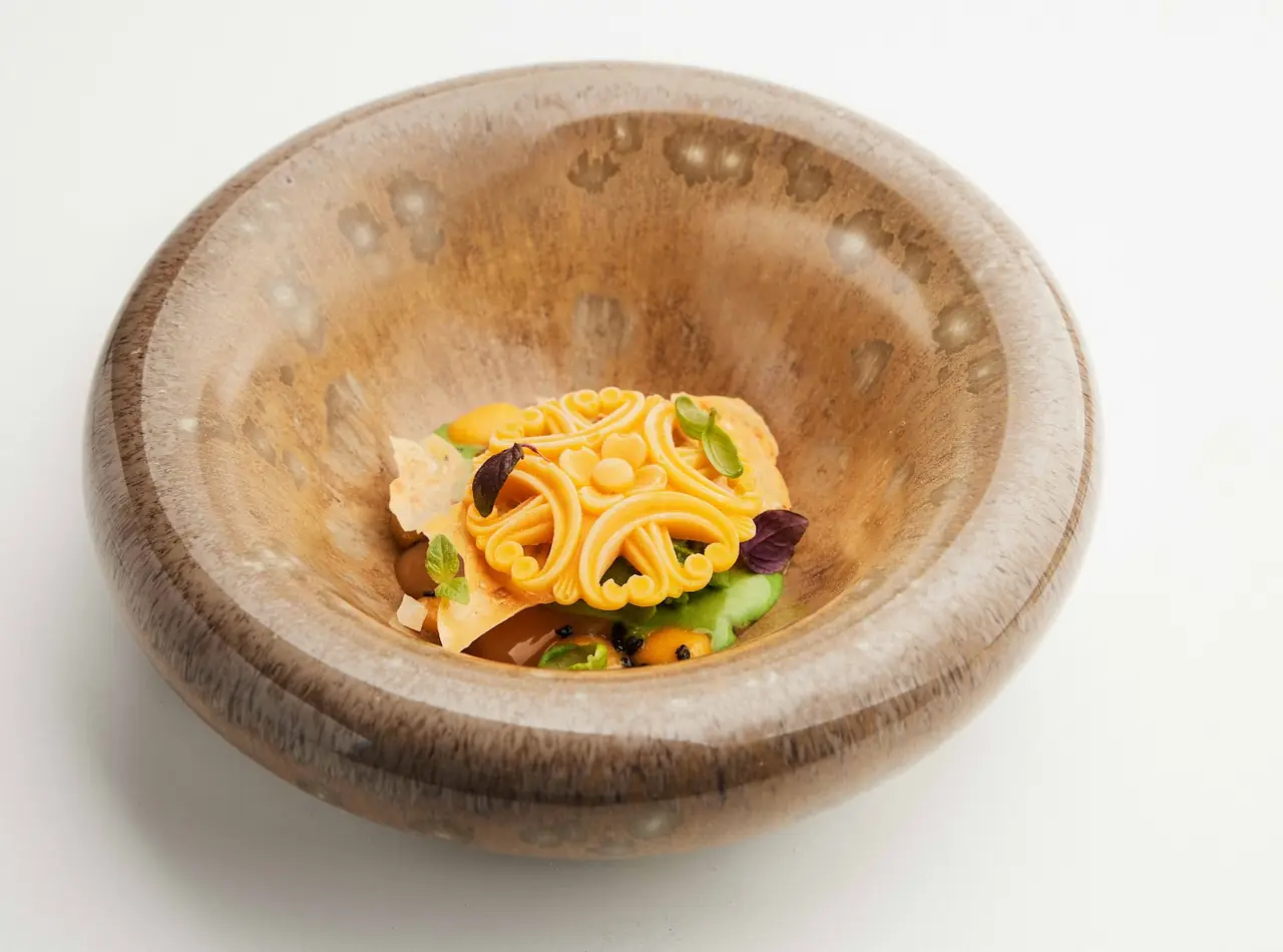 Avant-garde dish with ornate orange lattice piece over greens and citrus segments in deep wooden bowl