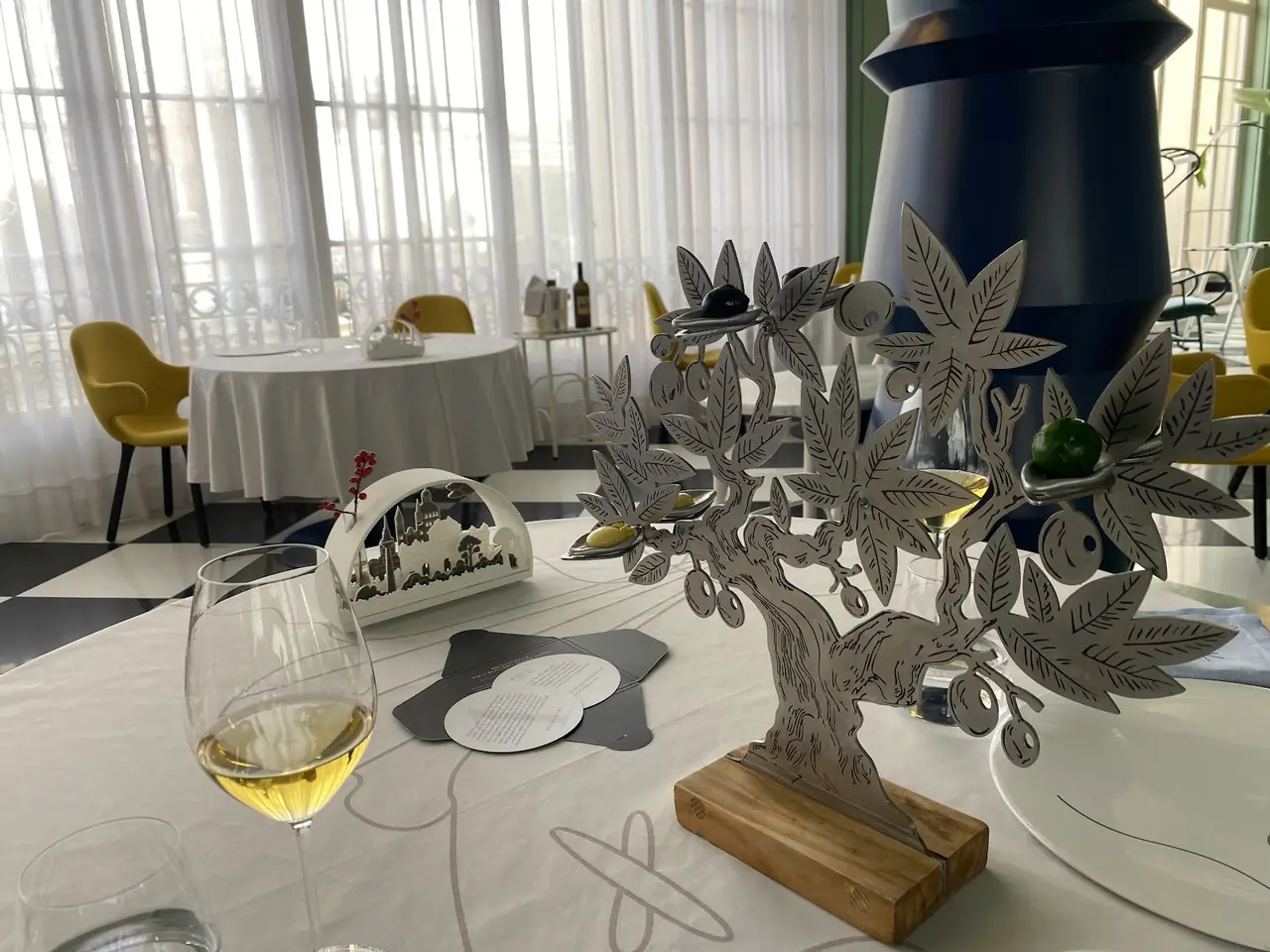 Glass of poured white wine on set table beside decorative olive tree centerpiece in dining room