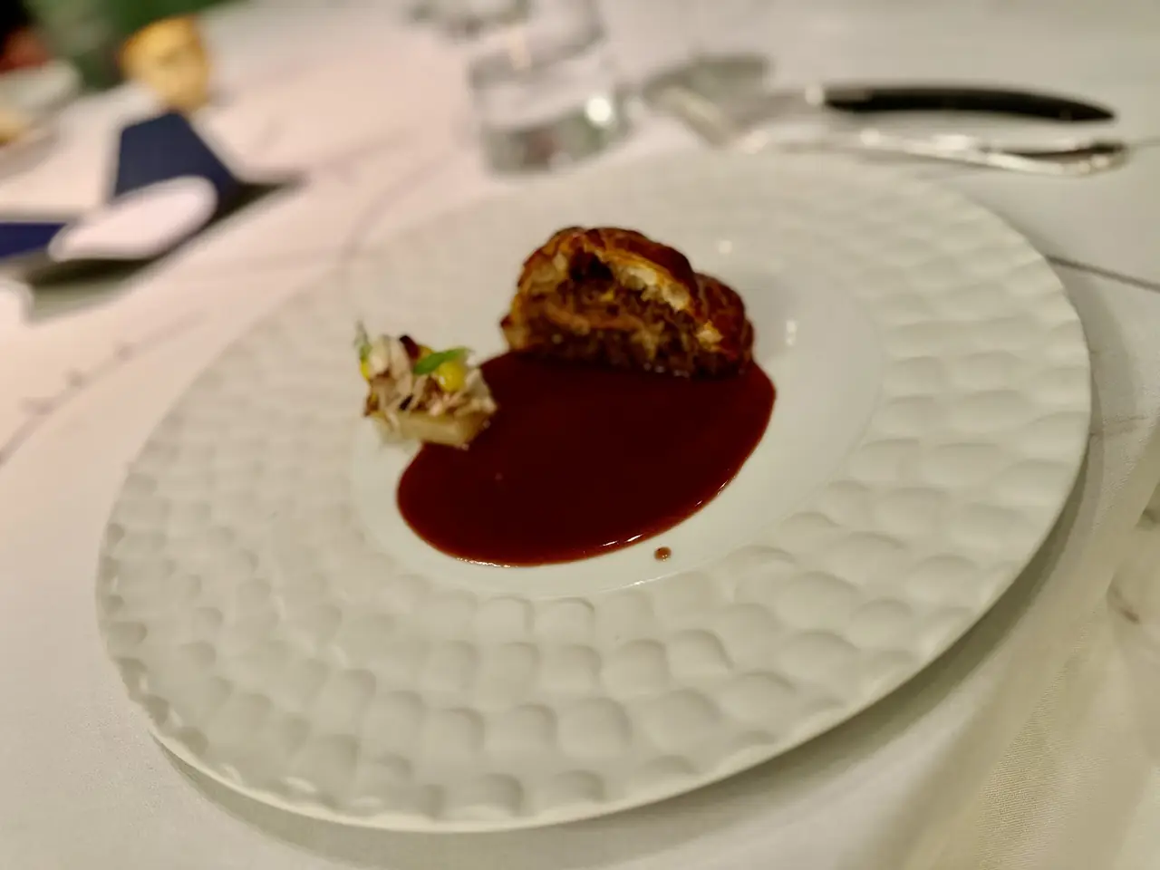 Small plated dish with pastry-wrapped bite in dark red sauce on textured white plate