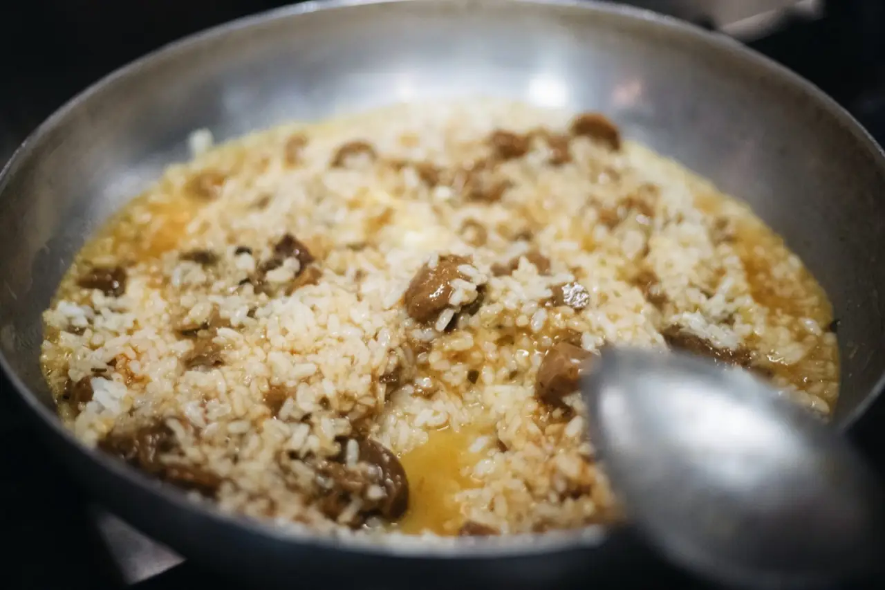 Rice cooked with mushrooms in a broth being stirred with a spoon in a steel pan