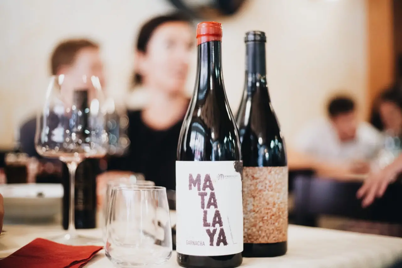 Two red-wine bottles including a Matalaya Garnacha and empty stemmed glasses on a white-clothed dining table, diners blurred behind