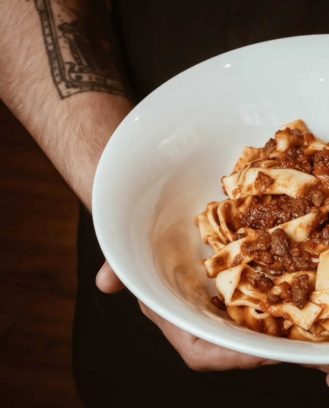 A hand holding a white bowl of wide pasta ribbons in a meat sauce
