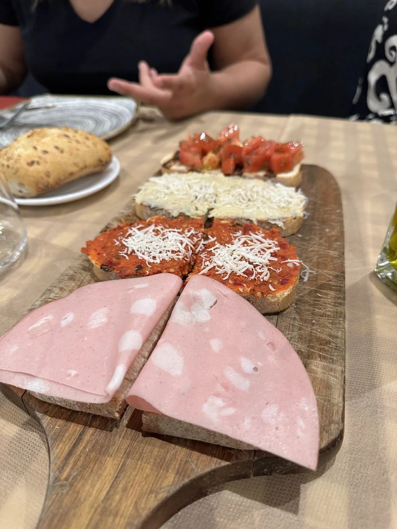 A wooden board with slices of pink mortadella, tomato-topped toast and bread
