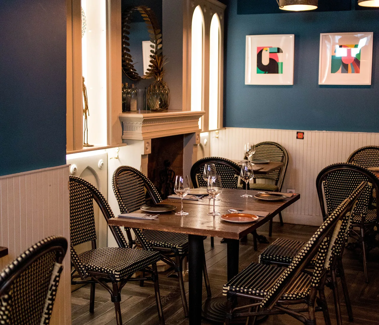 Dining room with deep teal walls, white panelling, black-and-cream bistro chairs around wooden tables set with wine glasses, and two bright graphic toucan prints on the wall