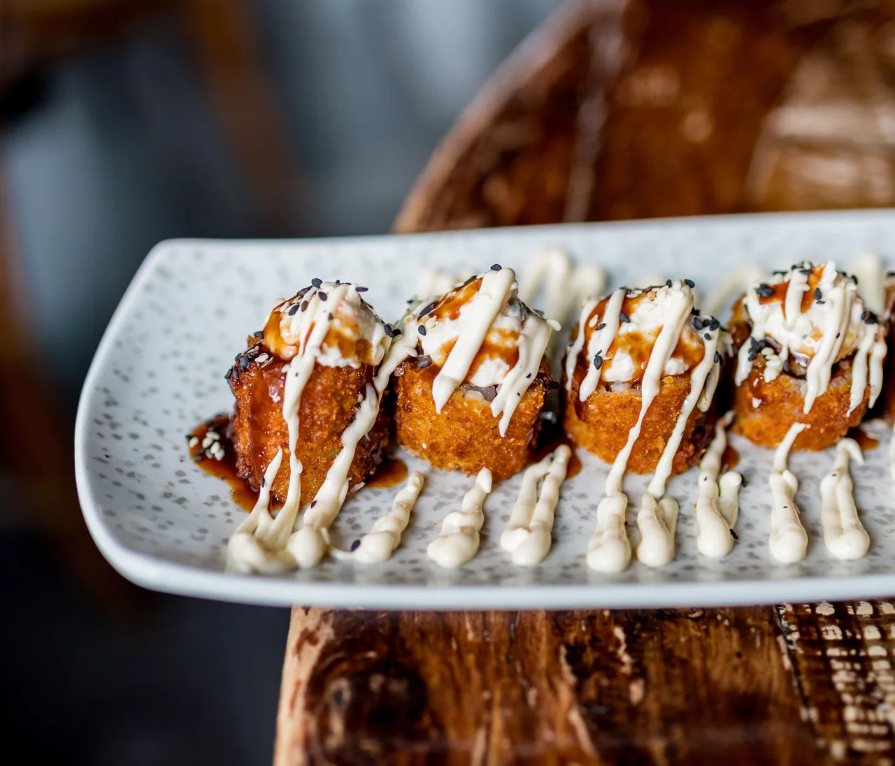 A row of deep-fried sushi rolls on a speckled rectangular plate, drizzled with pale mayonnaise and dark sauce and scattered with black and white sesame seeds