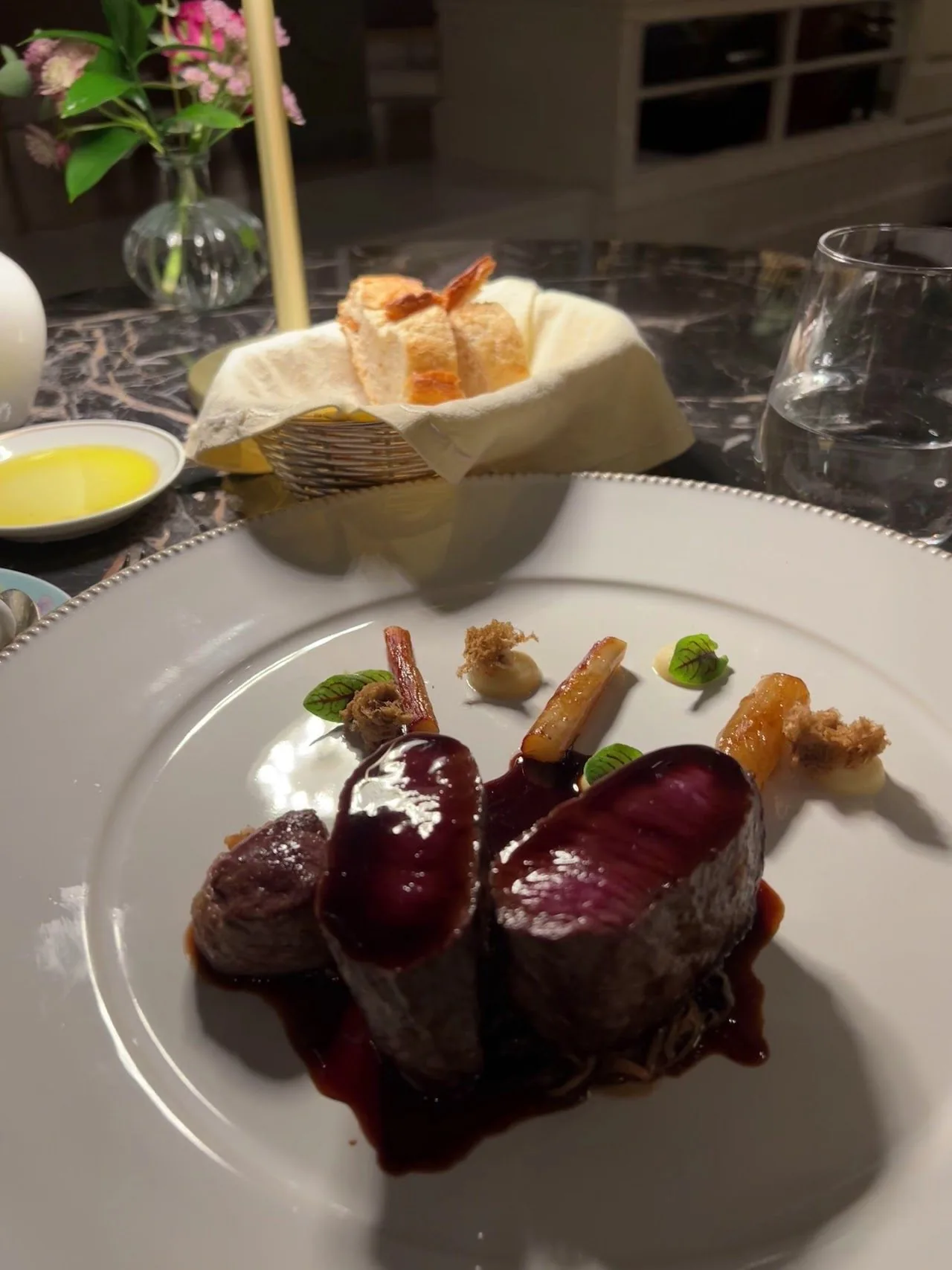 Slices of dark glazed meat with small vegetables and sauce on a large white plate, with a basket of bread behind