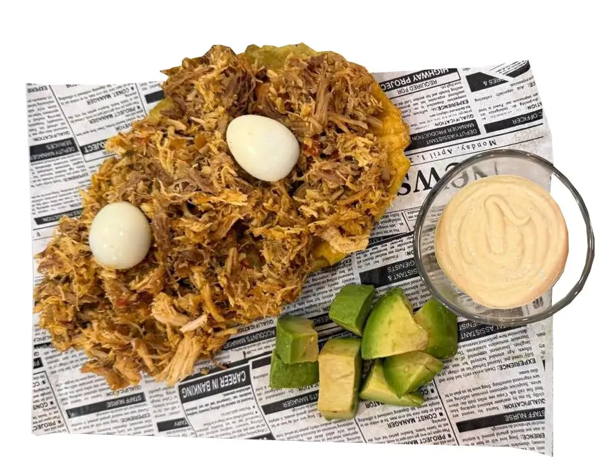 A fried green-plantain patacon topped with shredded meat and two quail eggs, with cubed avocado and a dish of pink sauce, on newspaper-print paper