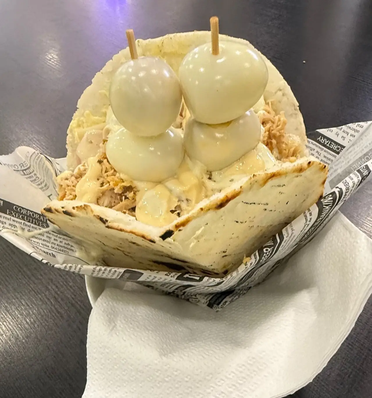 A grilled arepa filled with shredded chicken, cheese sauce and quail eggs on skewers, served in a newspaper-print paper tray
