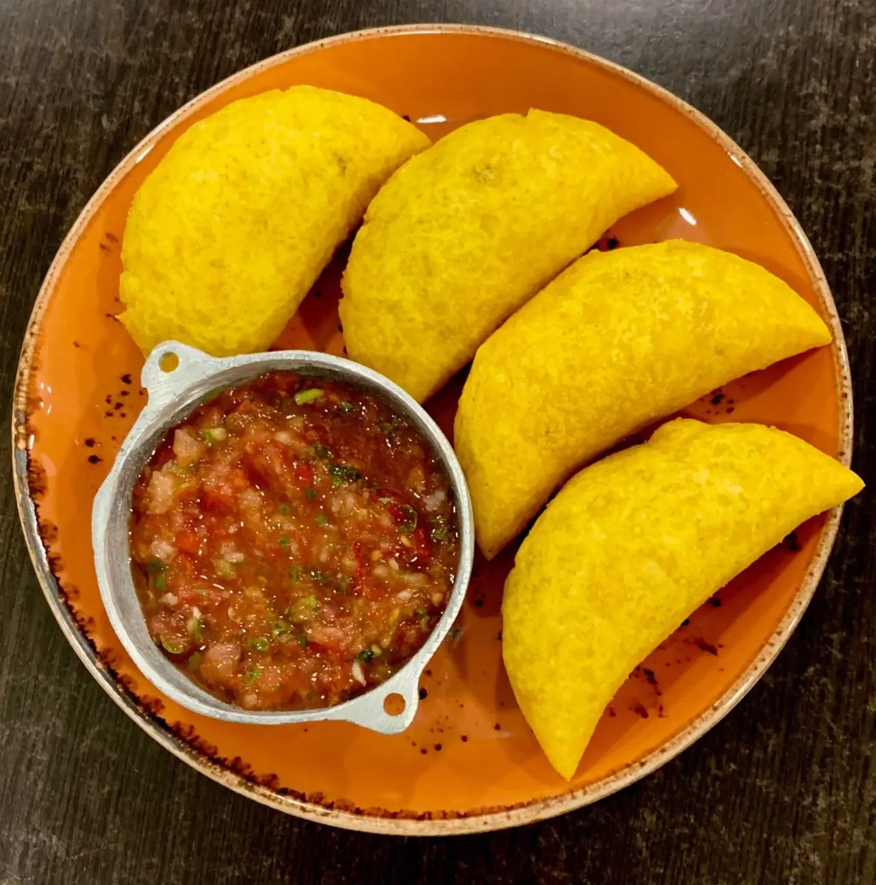 Four golden fried Colombian empanadas on an orange plate with a metal pot of chunky tomato-and-herb salsa