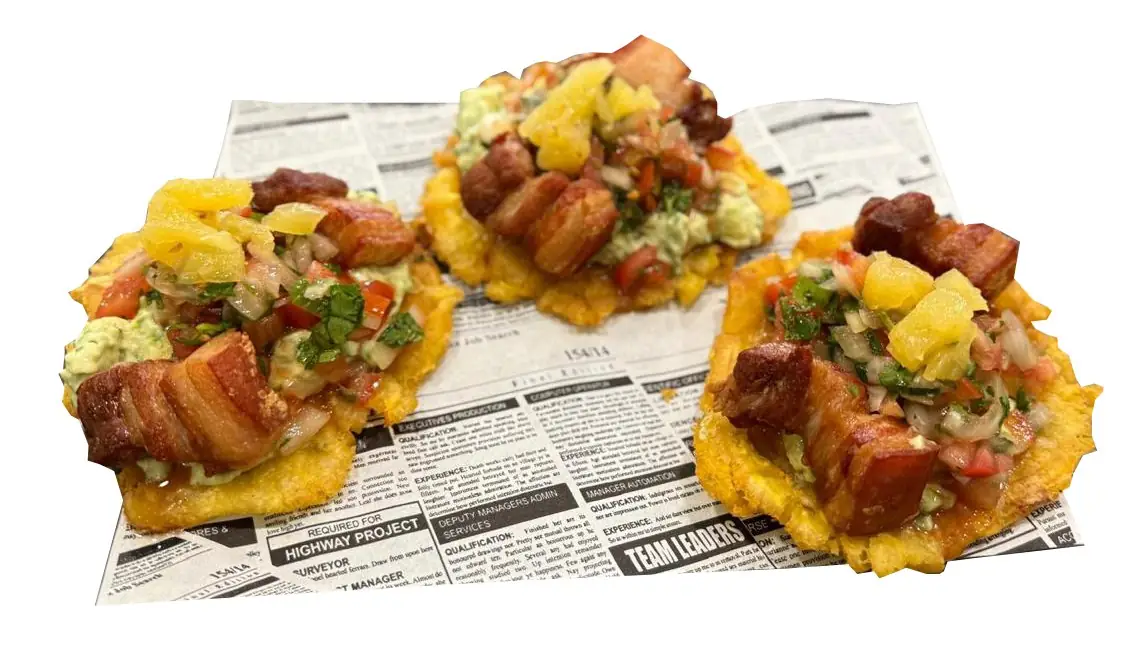 Three fried plantain patacones topped with pork belly, guacamole, pico de gallo and pineapple, on newspaper-print paper
