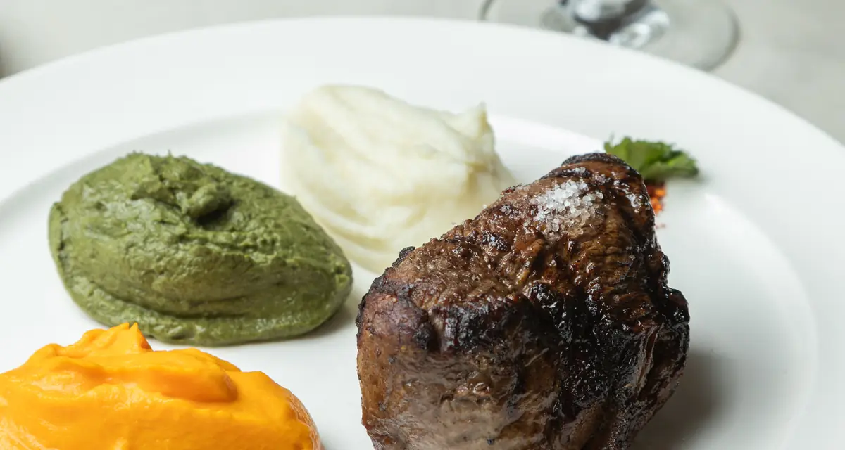 Grilled steak with green herb puree, mashed potato, and sweet potato puree on a white plate at Patagònia Beef & Wine