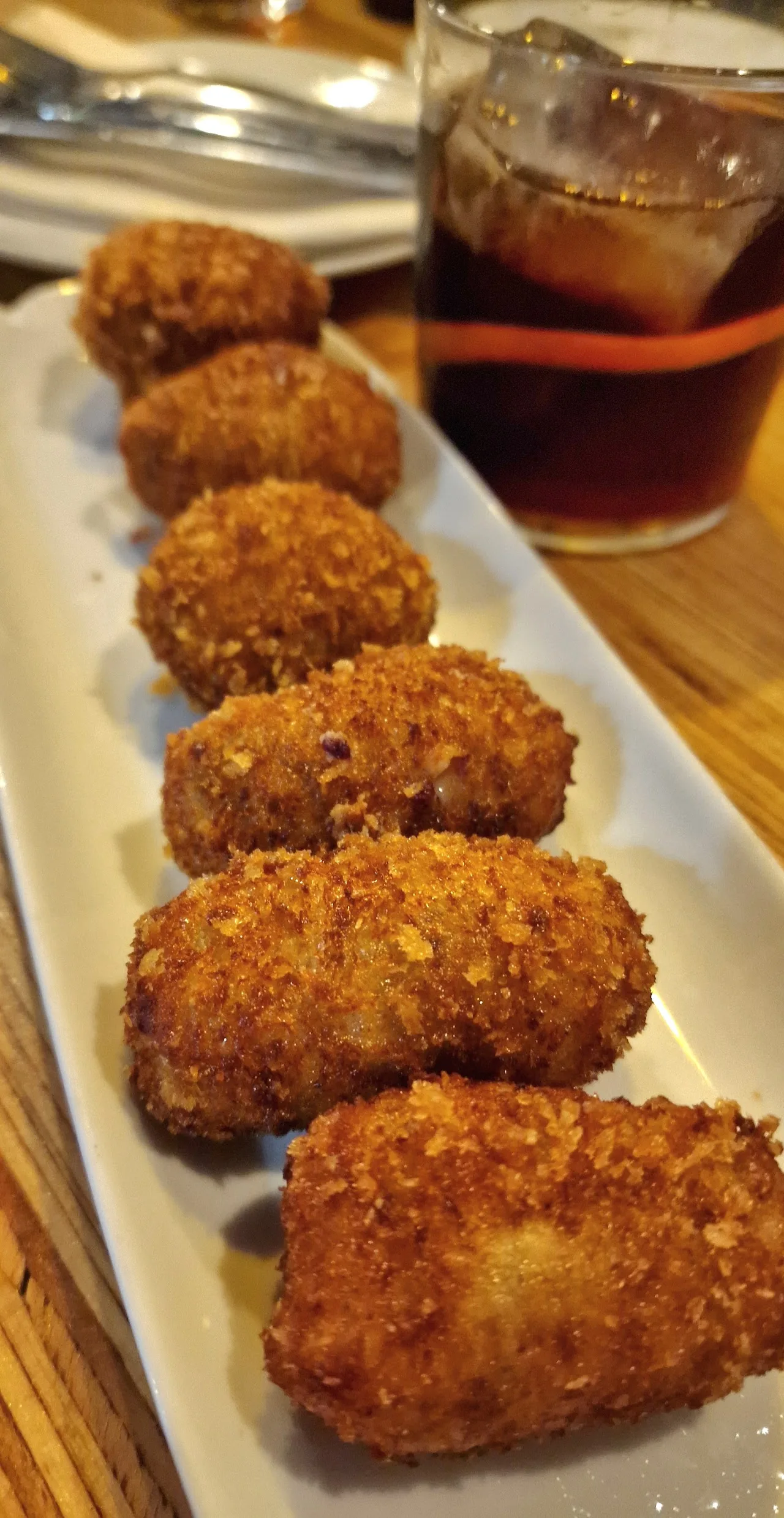 A row of breaded fried croquettes on a plate next to a glass of iced tea