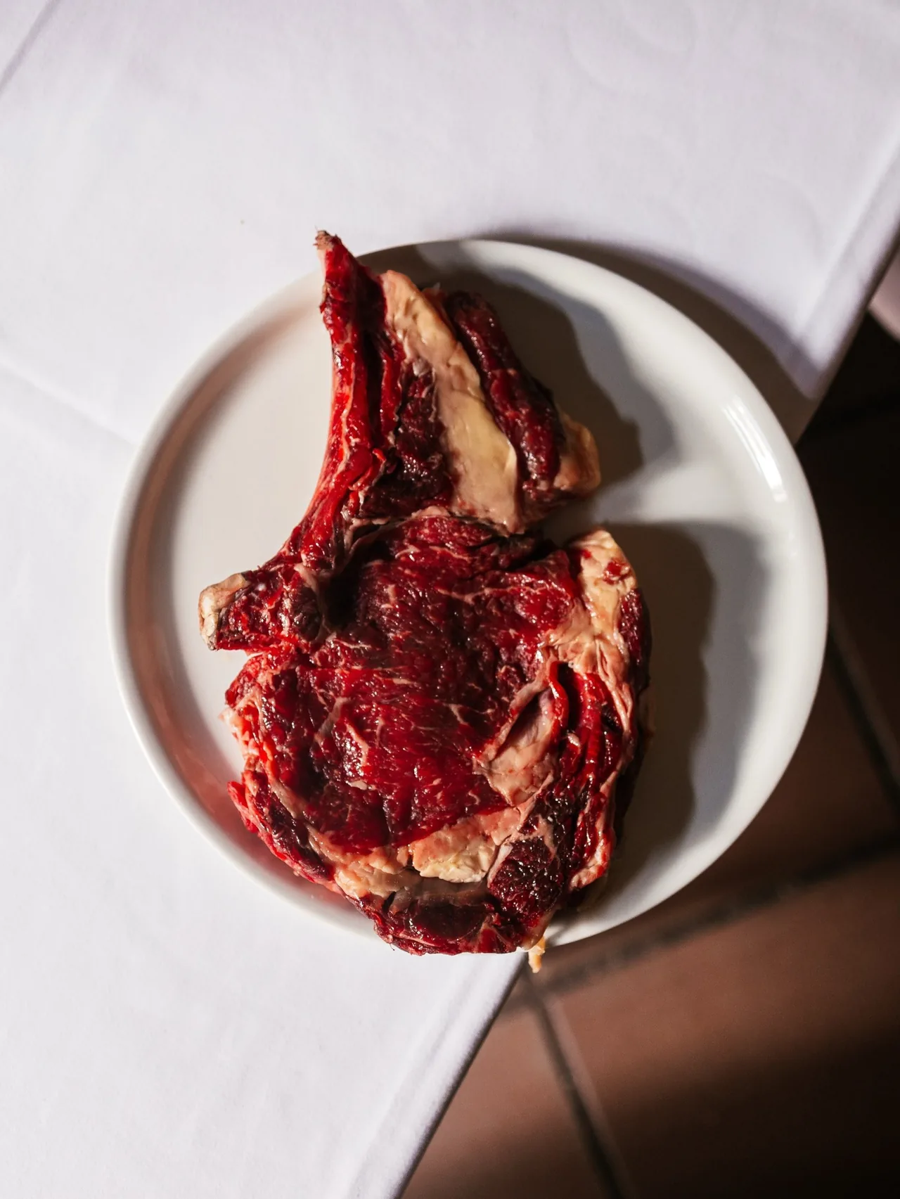 A raw bone-in beef chop on a white plate on a white tablecloth