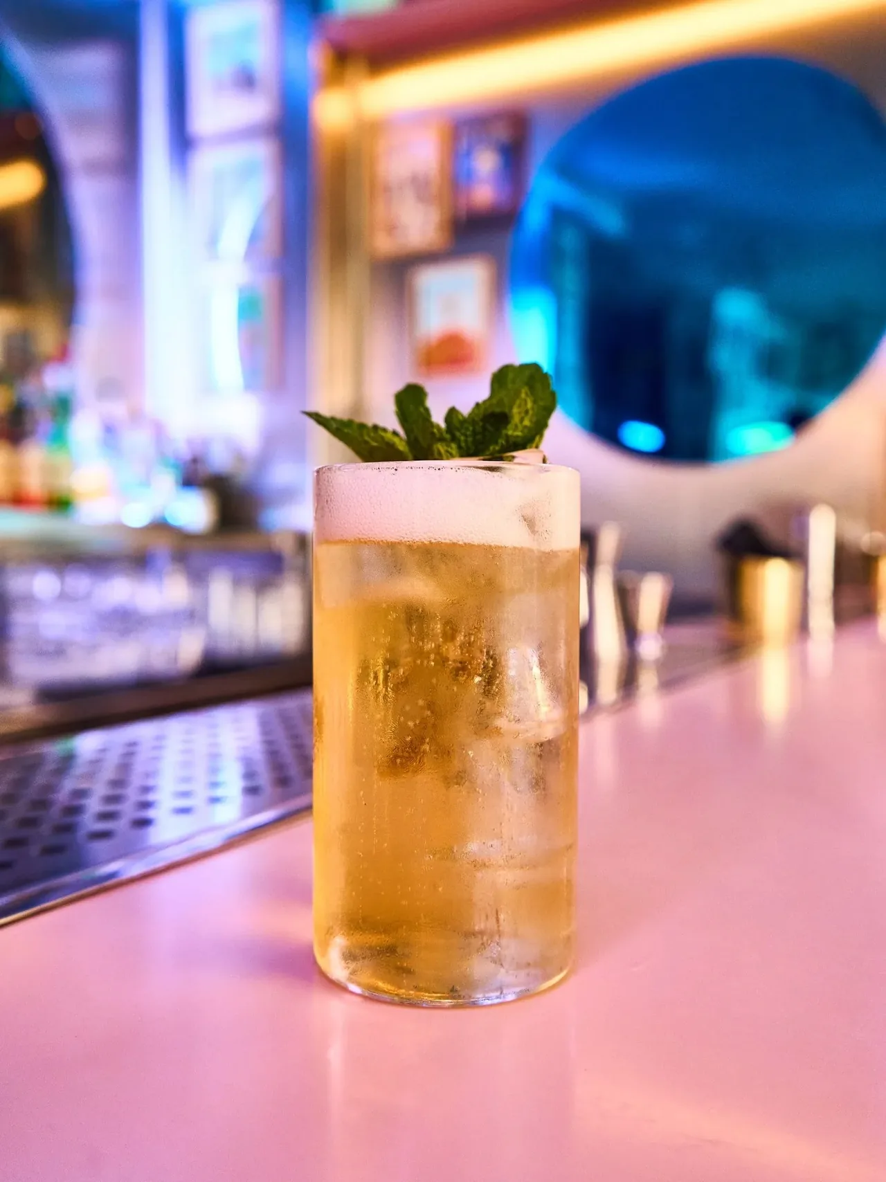 A tall golden cocktail with mint on a pink bar counter with a blurred bar behind it