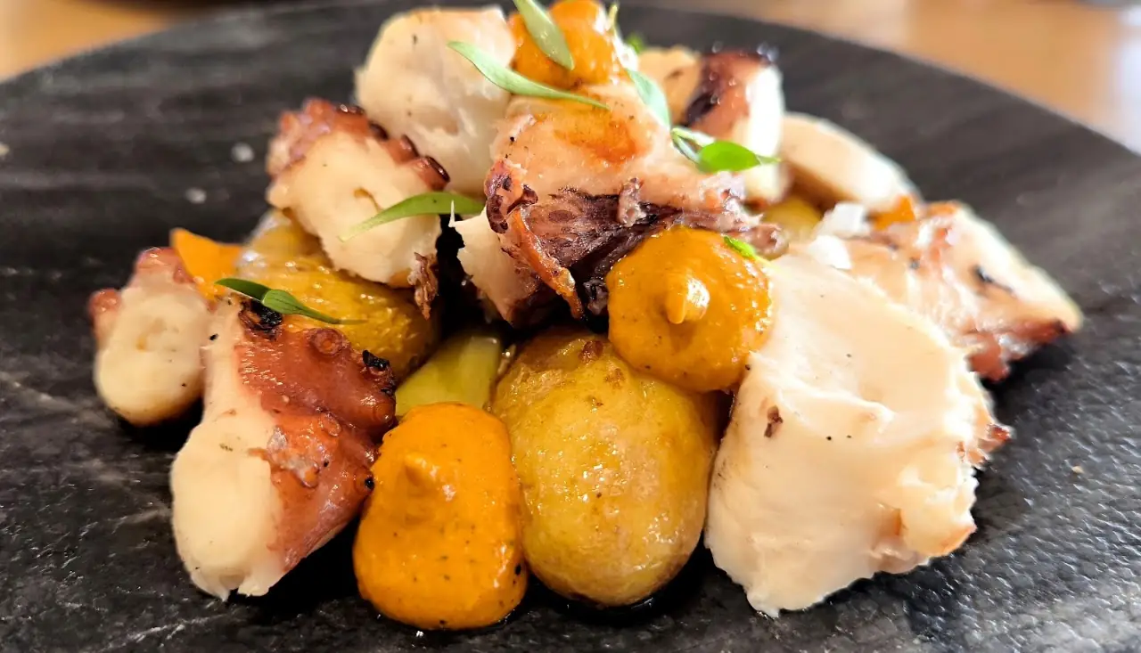 Grilled octopus with roasted baby potatoes and a creamy sauce on a dark plate.