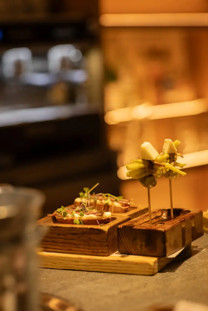 Pintxos on carved wooden holders on a bar top: a toast topped with meat and microgreens, and a skewered gilda of pickled pepper, anchovy, olive and cheese, warm blurred bar behind
