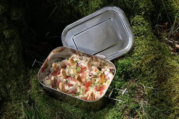 Creamy ensaladilla-style salad with diced tomato, radish, microgreens and crisp bread chips served in an open stainless-steel tin set on mossy ground